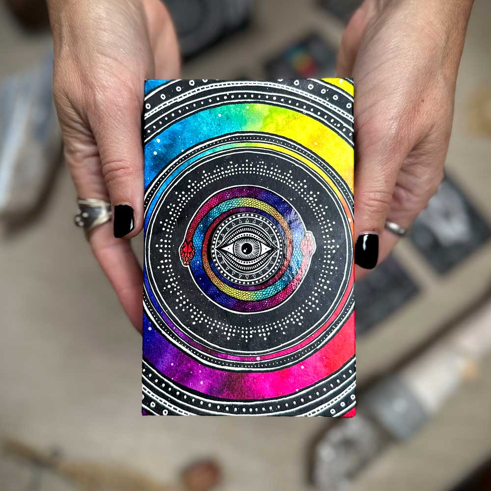 Hands holding a colorful Cosmic Visions Tarot notebook with concentric circles, dot patterns, and vibrant hues like blue, purple, red, and yellow centered around an eye design.