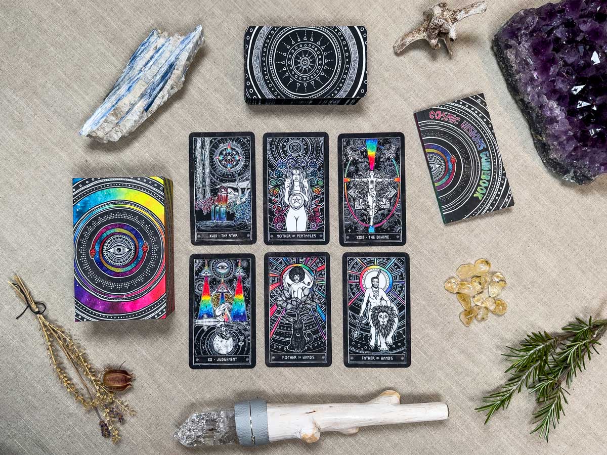 The Cosmic Visions Tarot spread adorns a beige tablecloth, surrounded by crystals, stones, and rosemary. The cards feature colorful, intricate designs with the deck at the top and crystals around this mystical setup.