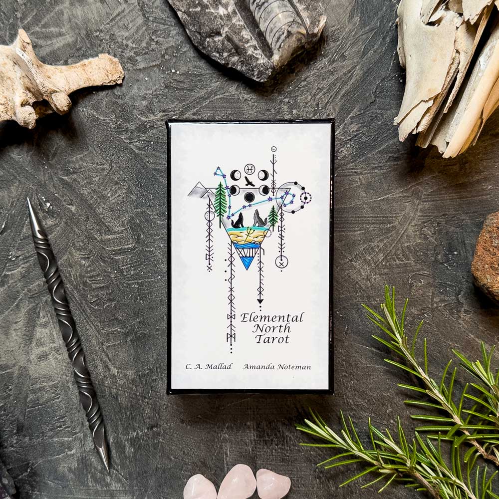 The Elemental North Tarot box rests elegantly on a dark textured surface, surrounded by assorted rocks, a twisted pen, rosemary sprigs, and small rose quartz stones. Its intricate geometric designs beg exploration of the mystical forces within.