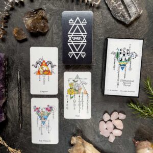 A tarot card spread on a dark surface includes the Elemental North Tarot cards—"Emperor," "Fool," and "8 of Pentacles"—with crystals, rocks, and a geometric, botanical-themed box.