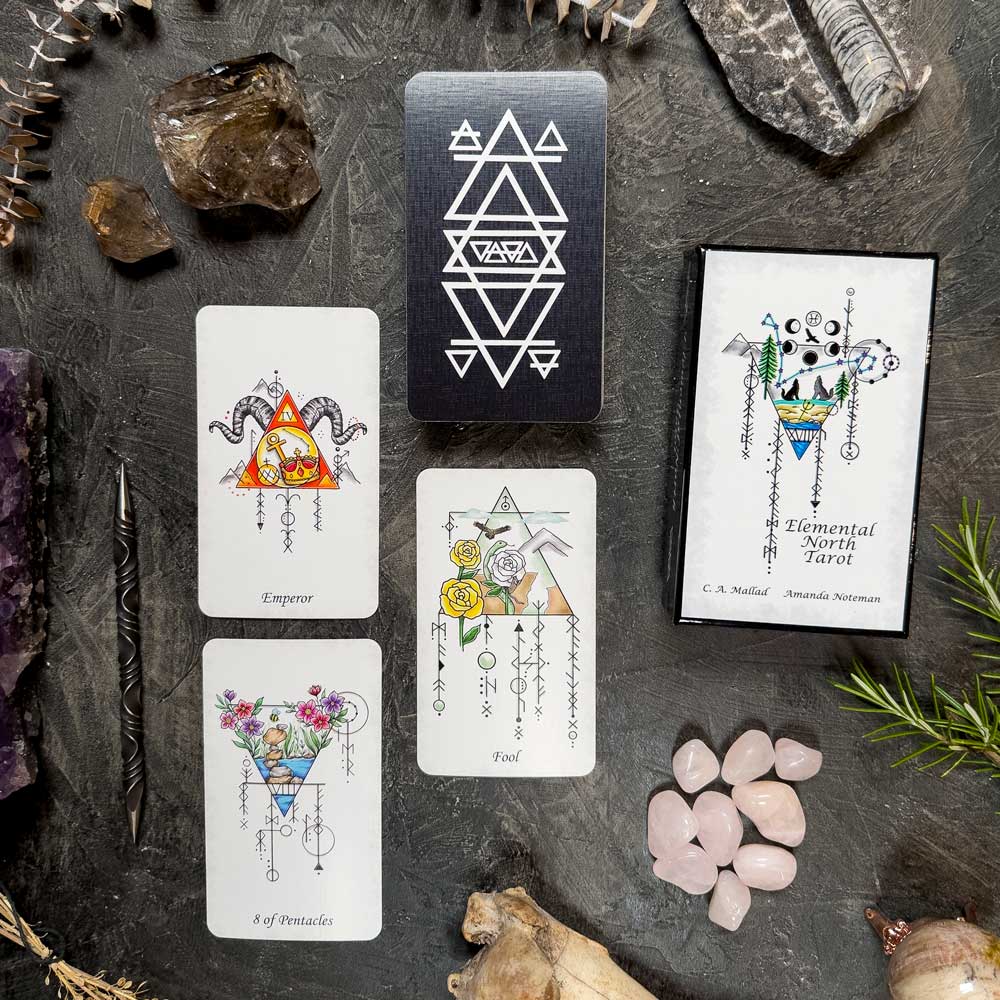 A tarot card spread on a dark surface includes the Elemental North Tarot cards—"Emperor," "Fool," and "8 of Pentacles"—with crystals, rocks, and a geometric, botanical-themed box.