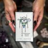 Hands adorned with rings and black nail polish hold the Elemental North Tarot deck, featuring abstract designs, mystical symbols, and a landscape within a triangle on its cover.