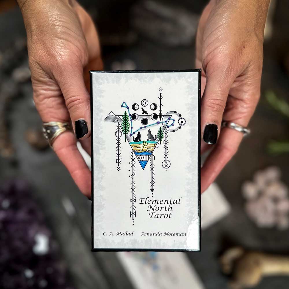 Hands adorned with rings and black nail polish hold the Elemental North Tarot deck, featuring abstract designs, mystical symbols, and a landscape within a triangle on its cover.