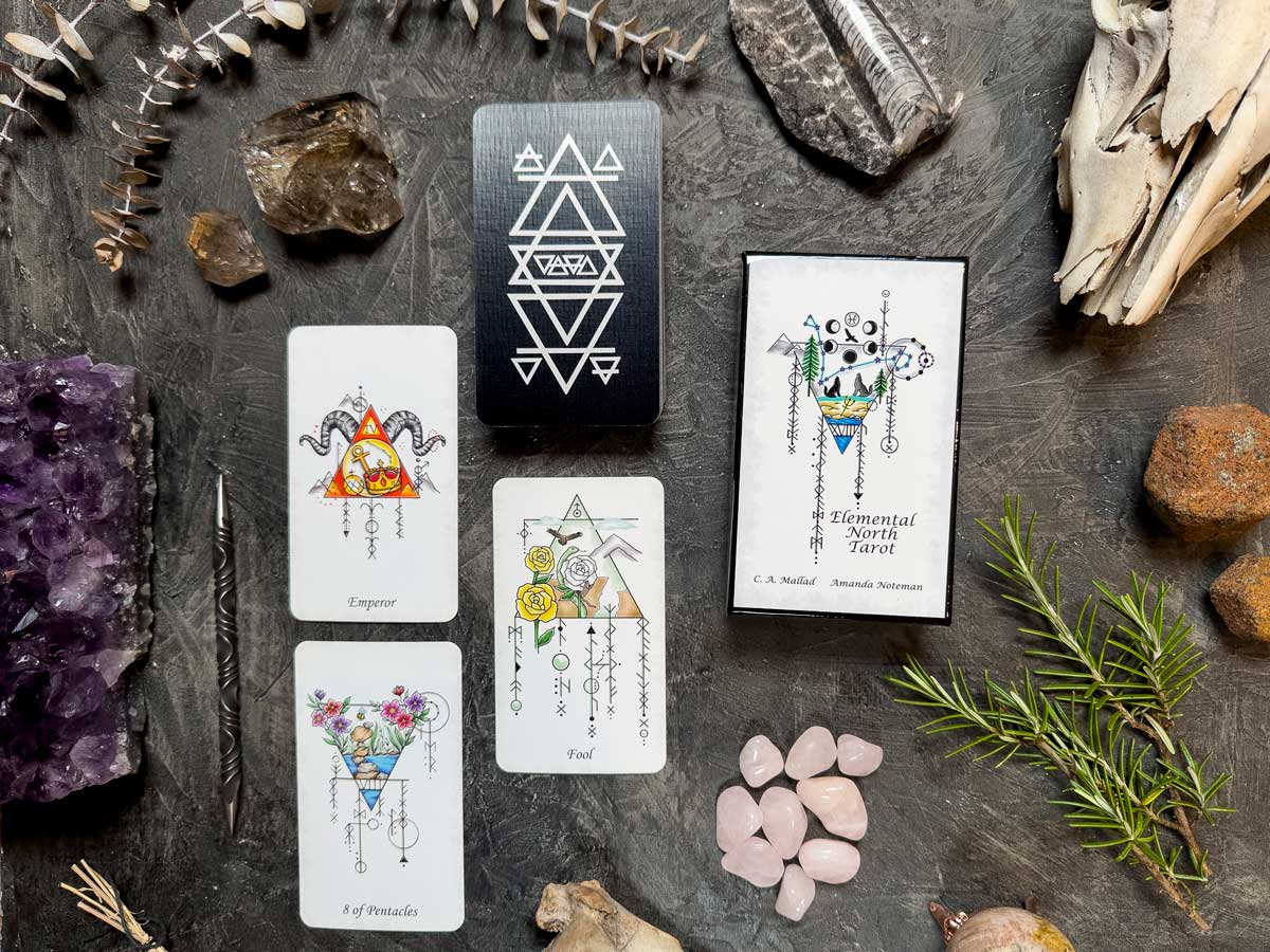 The Elemental North Tarot features a set with three cards, including the Emperor and Fool. Each card boasts geometric and floral designs. The deck box exhibits abstract patterns, surrounded by crystals, stones, and dried plants on a dark surface.