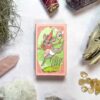 The image displays The Forest Enby Tarot deck beautifully arranged with crystals, herbs, and a skull on a white background. The tarot box features an illustration of a deer-like creature juggling.