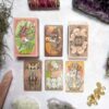 The Forest Enby Tarot set, featuring whimsical animal-themed cards, is beautifully displayed on a surface. Surrounded by crystals, dried herbs, and stones, it creates an enchanting scene that invites exploration.