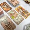 The Forest Enby Tarot spread showcases its intricately illustrated, colorful cards with mystical and animal themes on a white surface. In the background, you can see a stack of these enchanting cards alongside a skull decoration.