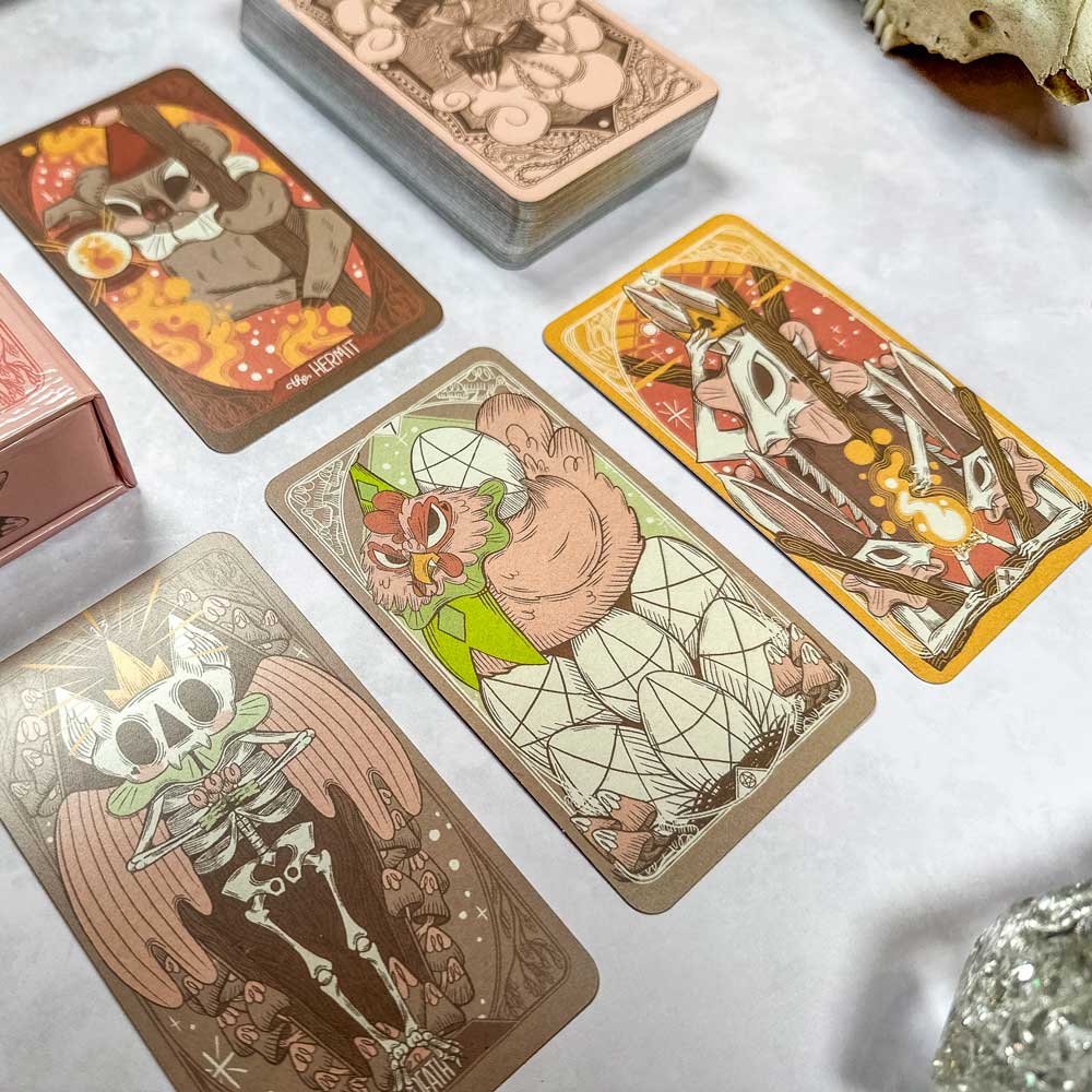 The Forest Enby Tarot spread showcases its intricately illustrated, colorful cards with mystical and animal themes on a white surface. In the background, you can see a stack of these enchanting cards alongside a skull decoration.