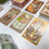Four illustrated tarot cards from The Forest Enby Tarot are displayed, featuring animal and mystical designs. A stack of similar cards is partially visible on the right next to a crystal, with a light-colored background.