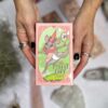 Hands hold "The Forest Enby Tarot," featuring whimsical art of an embroidered masked deer with a scepter. Blurred textured objects in the background create an enchanting tapestry, enhancing the deck's mystical allure.