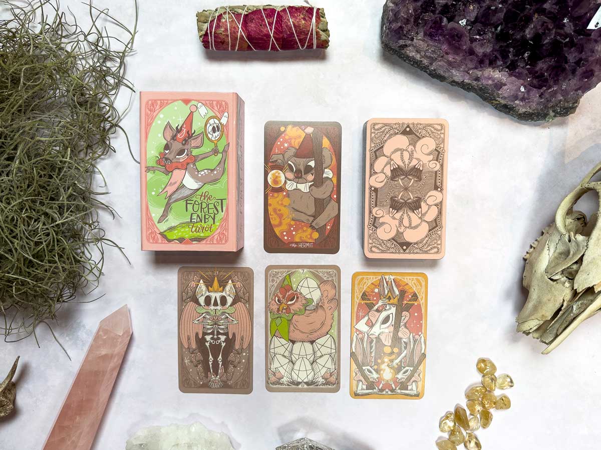 Spread across the table, "The Forest Enby Tarot" cards reveal whimsical animal characters surrounded by crystals, a sage bundle, and dried moss.