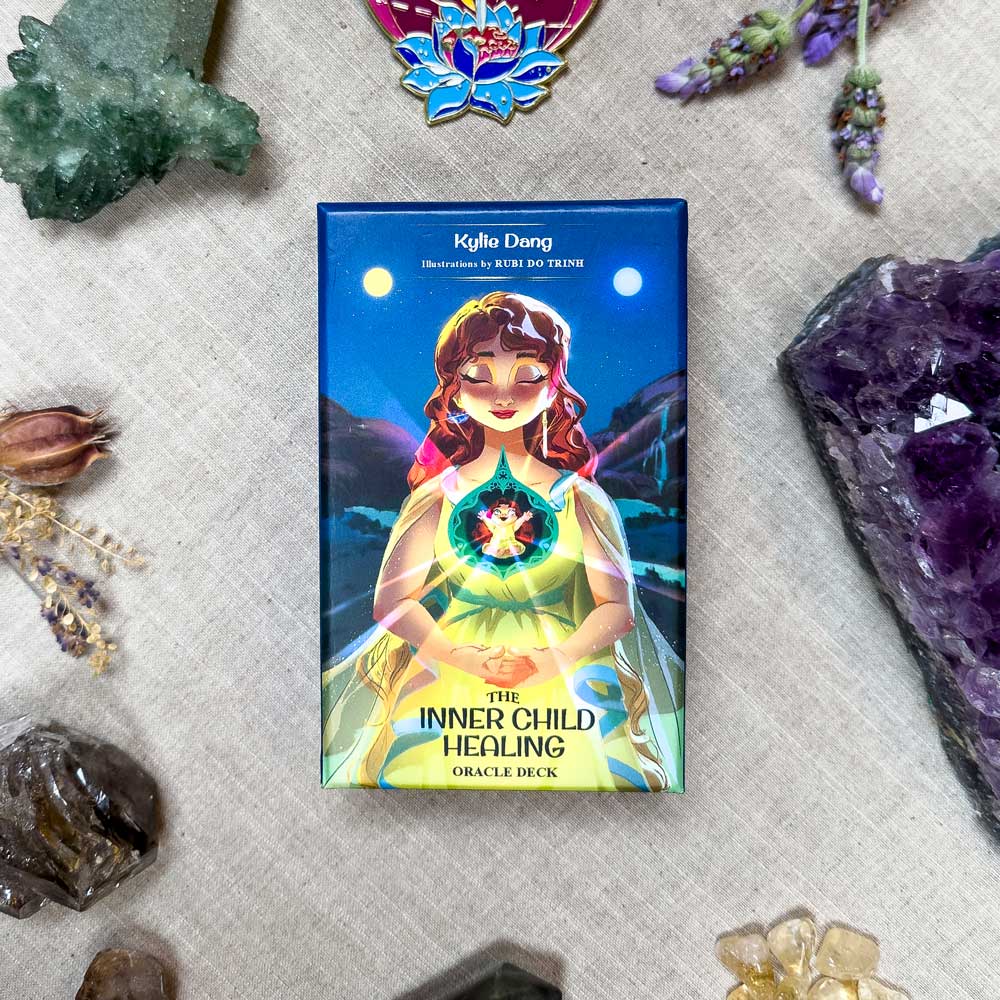 The Inner Child Healing Oracle sits on a table, its cover illustrated with a girl and energy sphere. Crystals, dried flowers, and a butterfly pin elegantly surround the deck, capturing the essence of inner child healing.
