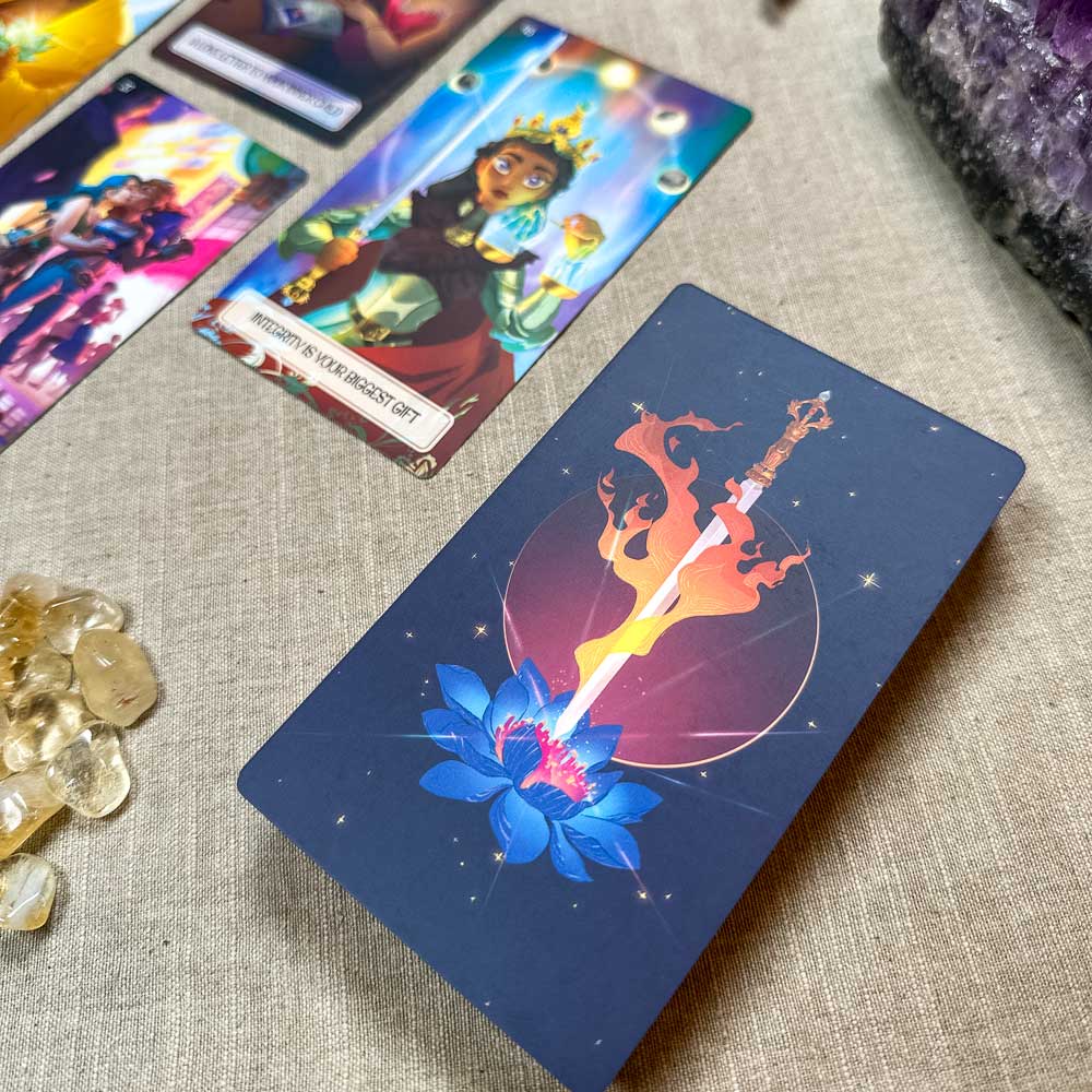 A tarot spread is laid out on a cloth, showcasing colorful cards. Prominent is The Inner Child Healing Oracle card illustrating a sword piercing a blue lotus against a cosmic background. Nearby, small translucent gemstones and an amethyst crystal softly shimmer.