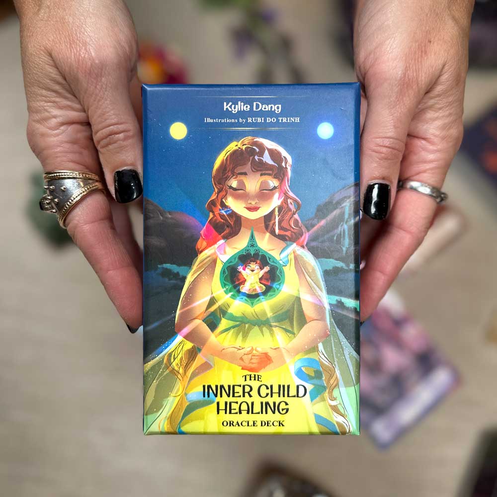 A person holds "The Inner Child Healing Oracle," featuring vibrant cover art of a woman with glowing elements by Rubi Qi Trinh, complementing their sleek black nails.