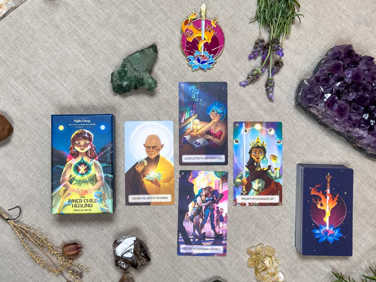 An oracle card spread sits on a cloth with crystals, dried herbs, and a pendant. Enhancing this magical scene is The Inner Child Healing Oracle deck, its cards and box decorated with vibrant illustrations.
