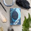 The Lubanko Tarot, in an illustrated blue and black box, is artistically arranged on a beige cloth with crystals, a dark stone, fern leaf, metal circle, and carved wooden wand.