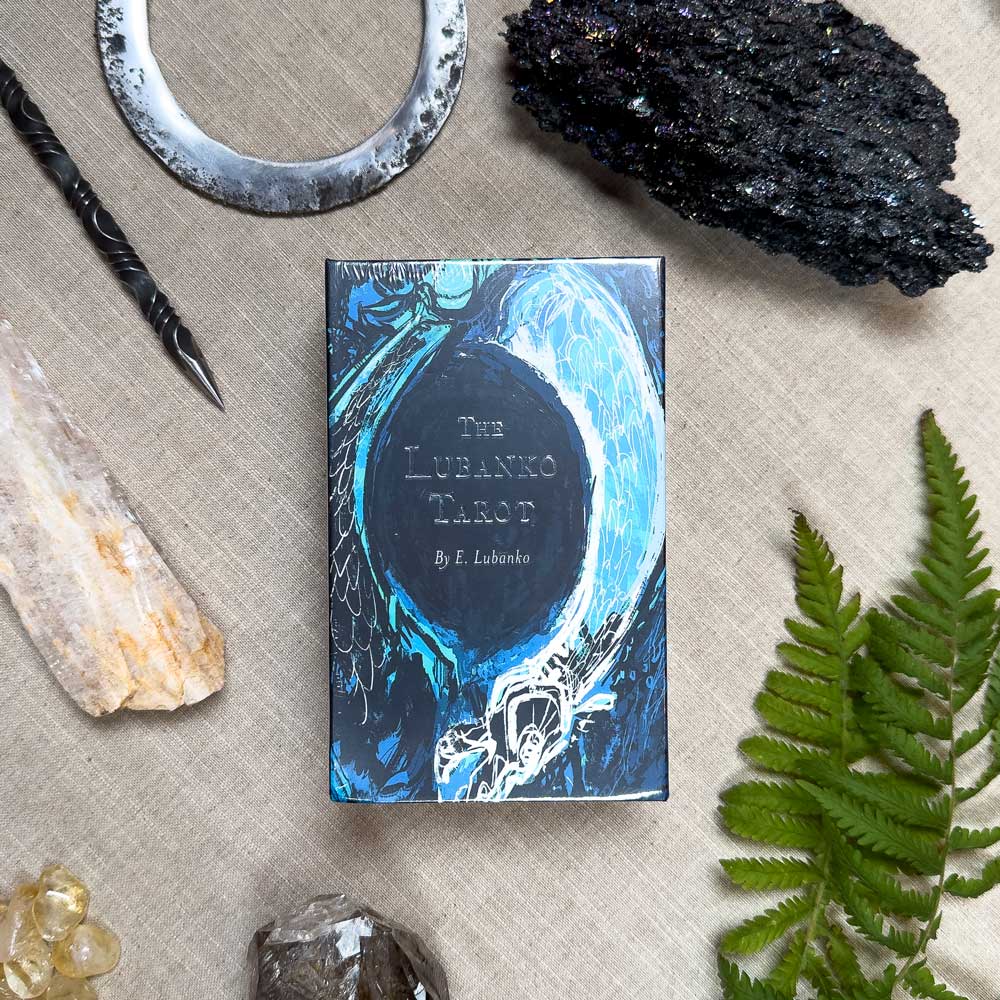 The Lubanko Tarot, in an illustrated blue and black box, is artistically arranged on a beige cloth with crystals, a dark stone, fern leaf, metal circle, and carved wooden wand.