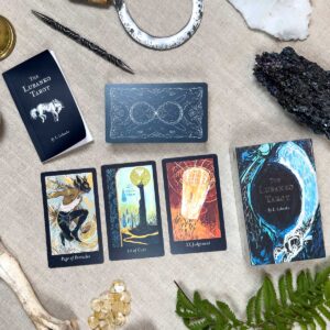 A tarot reading setup showcases three Lubanko Tarot cards: Page of Pentacles, 10 of Cups, and Judgment. Crystals, a fern, and guidebooks embellish the scene on a beige cloth backdrop.