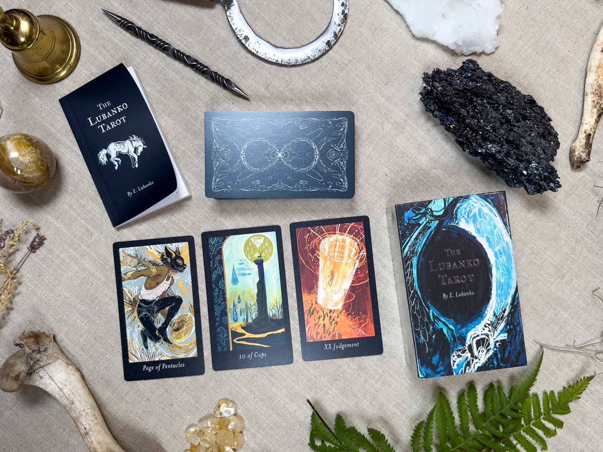 A textured surface hosts The Lubanko Tarot setup. Four cards with artistic abstract designs lie face up among crystals, horns, a brass object, fern leaves, and a tarot guidebook, radiating enigmatic beauty.
