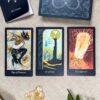 Three tarot cards from The Lubanko Tarot are laid out: Page of Pentacles with a leaping rabbit, 10 of Cups showing a tower and cups, and XX Judgment depicting a radiant angelic figure. Nearby, crystals glisten among lush greenery.