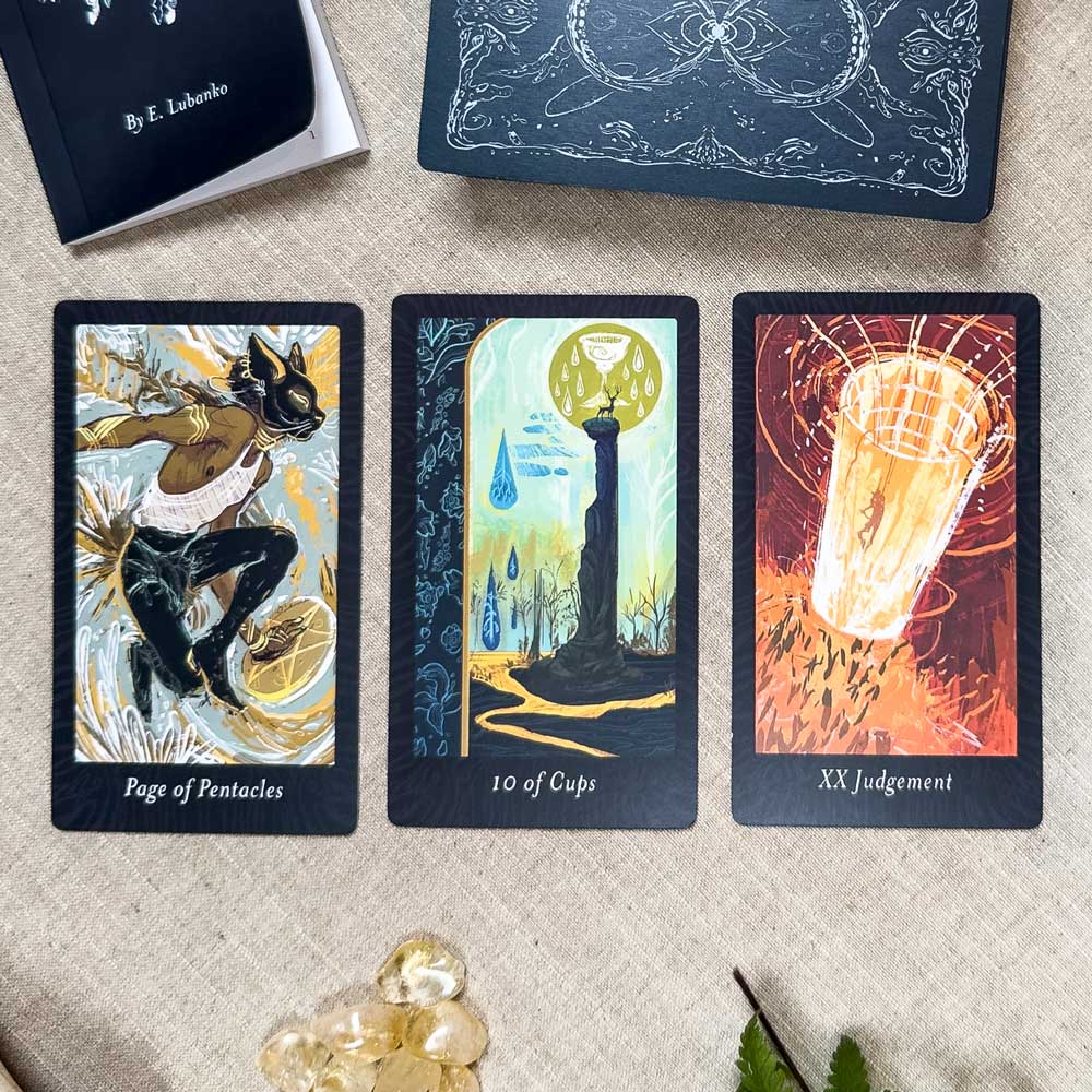 Three tarot cards from The Lubanko Tarot are laid out: Page of Pentacles with a leaping rabbit, 10 of Cups showing a tower and cups, and XX Judgment depicting a radiant angelic figure. Nearby, crystals glisten among lush greenery.