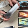 Close-up of hands with black nail polish holding The Lubanko Tarot deck with shimmering silver edges. Several illustrated cards from the deck are spread out on a table next to a crystal and guidebook.