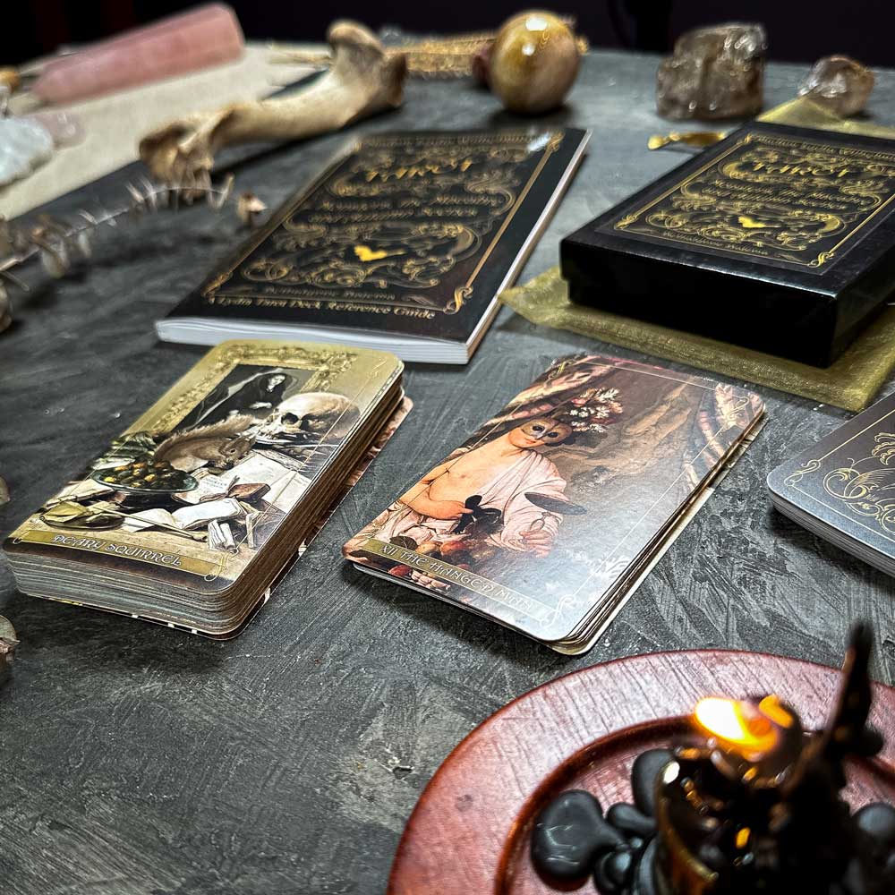 A mystical scene with Madam Lydia Wilhelmina's Tarot features ornate tarot cards and a deck showing a cloaked figure. Crystals, a carved wooden coaster, and a small candle enhance the ambiance on a textured dark surface.