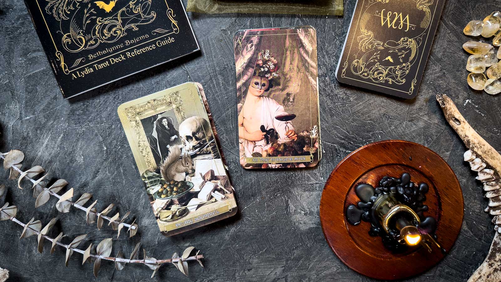 A decorative setup with Madam Lydia Wilhelmina's Tarot features Victorian-themed cards on a dark surface, accompanied by an open guidebook, black candle wax, crystals, a bone, an ink bottle, and dried foliage for a mysterious vintage vibe.