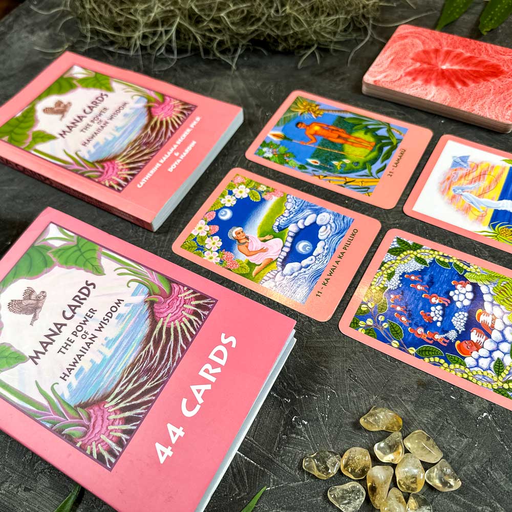 On a table, a set of Mana Cards displaying Hawaiian wisdom with vibrant illustrations is arranged alongside six cards, a guidebook, and small gemstones, all encircled by natural decor.