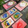 A set of Mana Cards is laid out, featuring six Hawaiian-themed oracle cards with vibrant illustrations of nature and mythology. Nearby, a box and guidebook accompany scattered stones and a leafy plant, creating a serene scene.