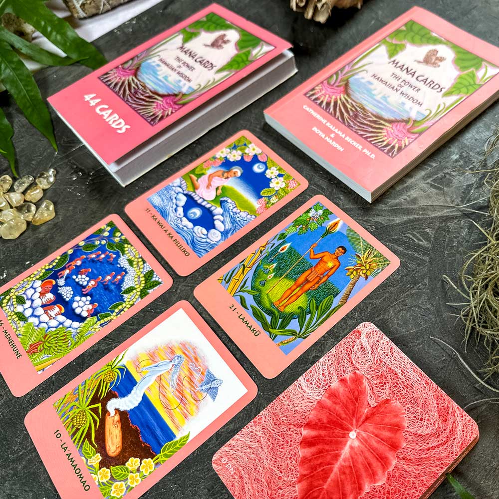 A set of Mana Cards is laid out, featuring six Hawaiian-themed oracle cards with vibrant illustrations of nature and mythology. Nearby, a box and guidebook accompany scattered stones and a leafy plant, creating a serene scene.