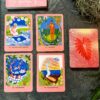 Five oracle cards, featuring the vivid Mana Cards, adorn a dark surface. Four display colorful illustrations labeled "Ka Wai A Ka Pililiko," "Lamakū," "Menehune," and "La ʻAmaʻamao." The fifth card lies face down with a striking red leaf design.