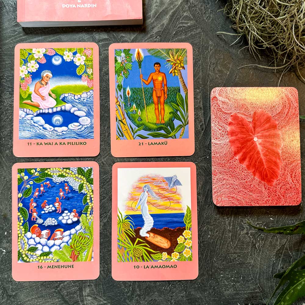 Five oracle cards, featuring the vivid Mana Cards, adorn a dark surface. Four display colorful illustrations labeled "Ka Wai A Ka Pililiko," "Lamakū," "Menehune," and "La ʻAmaʻamao." The fifth card lies face down with a striking red leaf design.