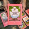 Two hands hold a book titled "Mana Cards," its pink cover featuring an owl and tropical plants, while beneath, the Mana Cards lie spread on a textured surface, inviting exploration of their rich insights and vibrant designs.