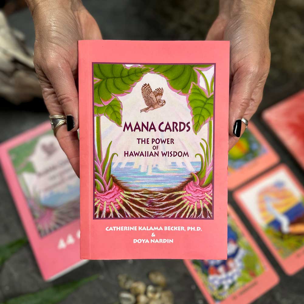 Two hands hold a book titled "Mana Cards," its pink cover featuring an owl and tropical plants, while beneath, the Mana Cards lie spread on a textured surface, inviting exploration of their rich insights and vibrant designs.
