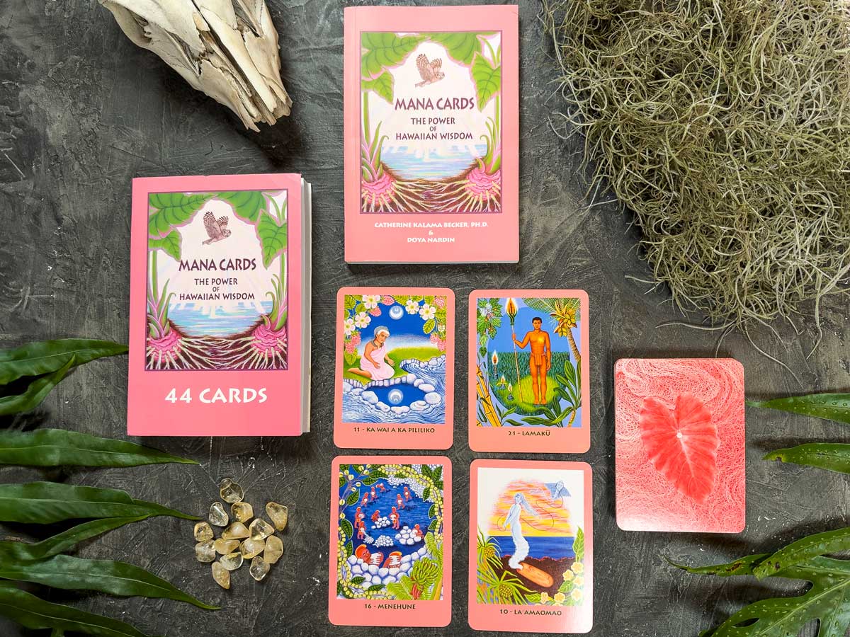 Mana Cards featuring Hawaiian themes like ocean landscapes and lush greenery are spread on a gray surface. The display includes a pink box, rocks, leaves, and cards with a red palm design.