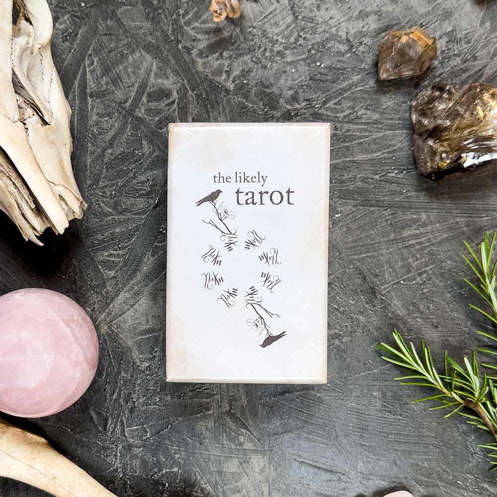 The Likely Tarot" box sits on a textured dark surface, surrounded by natural elements like a pink sphere, rosemary sprigs, driftwood, and smoky quartz crystals. This enchanting arrangement invites you to explore the mystical realm of The Likely Tarot.