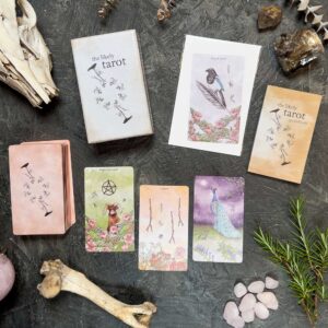The Likely Tarot cards, featuring animal and nature themes like birds, deer, and peacocks, are artistically displayed on a dark textured surface. The scene includes a tarot guidebook, crystals, and decorative animal skulls.
