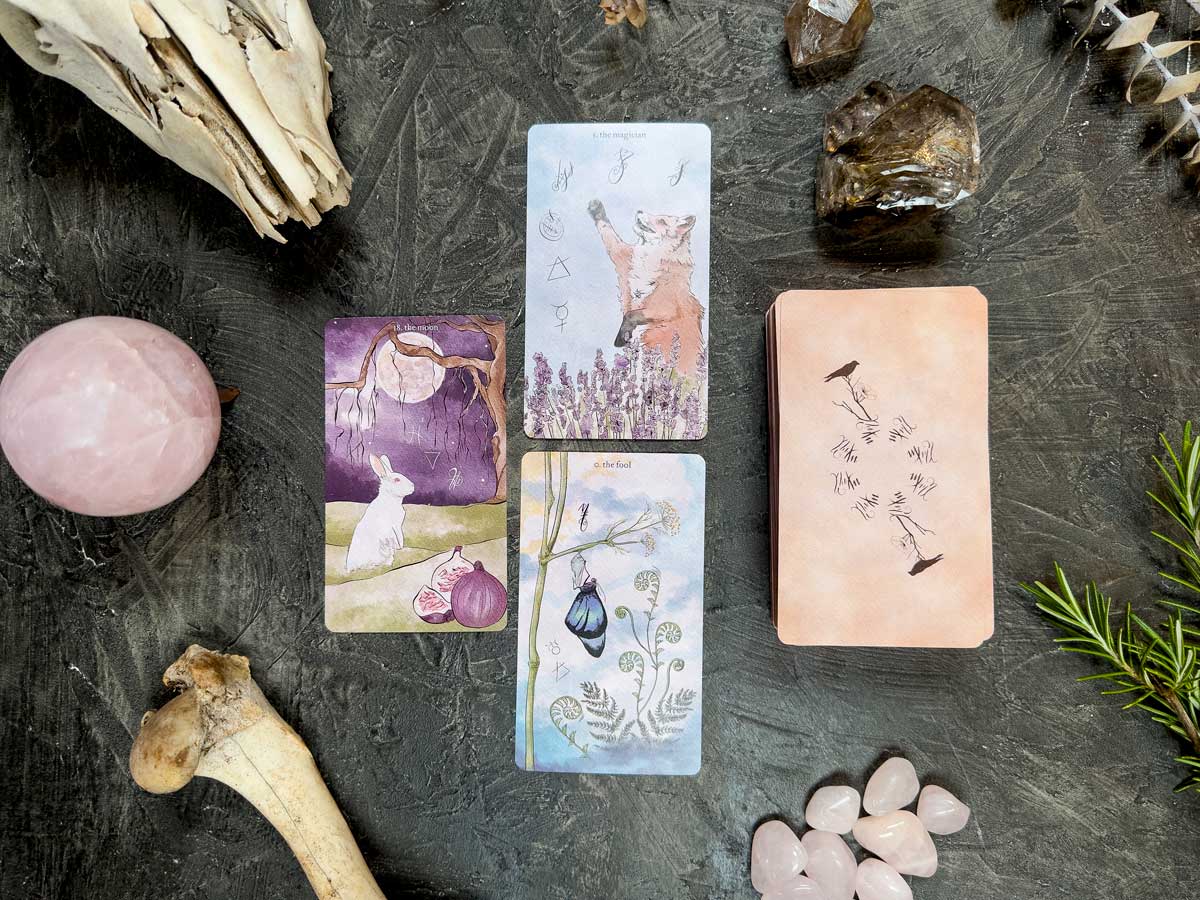A mystical arrangement features three Likely Tarot cards illustrating a rabbit, fox, and bird among crystals, bones, and greenery on a dark surface, with the deck nearby.