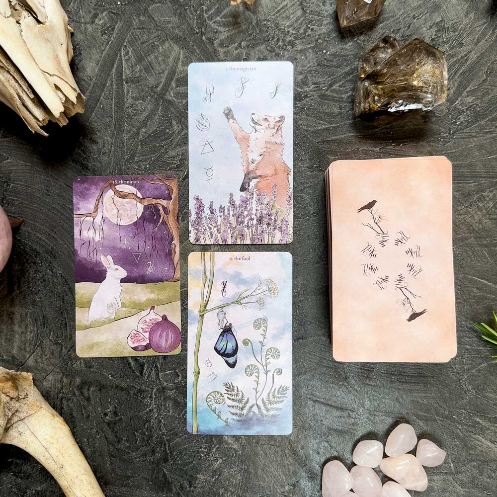 The dark surface showcases The Likely Tarot layout, decorated with crystals and bones. Three cards feature a fox, rabbit, and chrysalis. The deck rests close by, its back adorned with delicate illustrations for those intrigued by fate's mysterious tapestry.