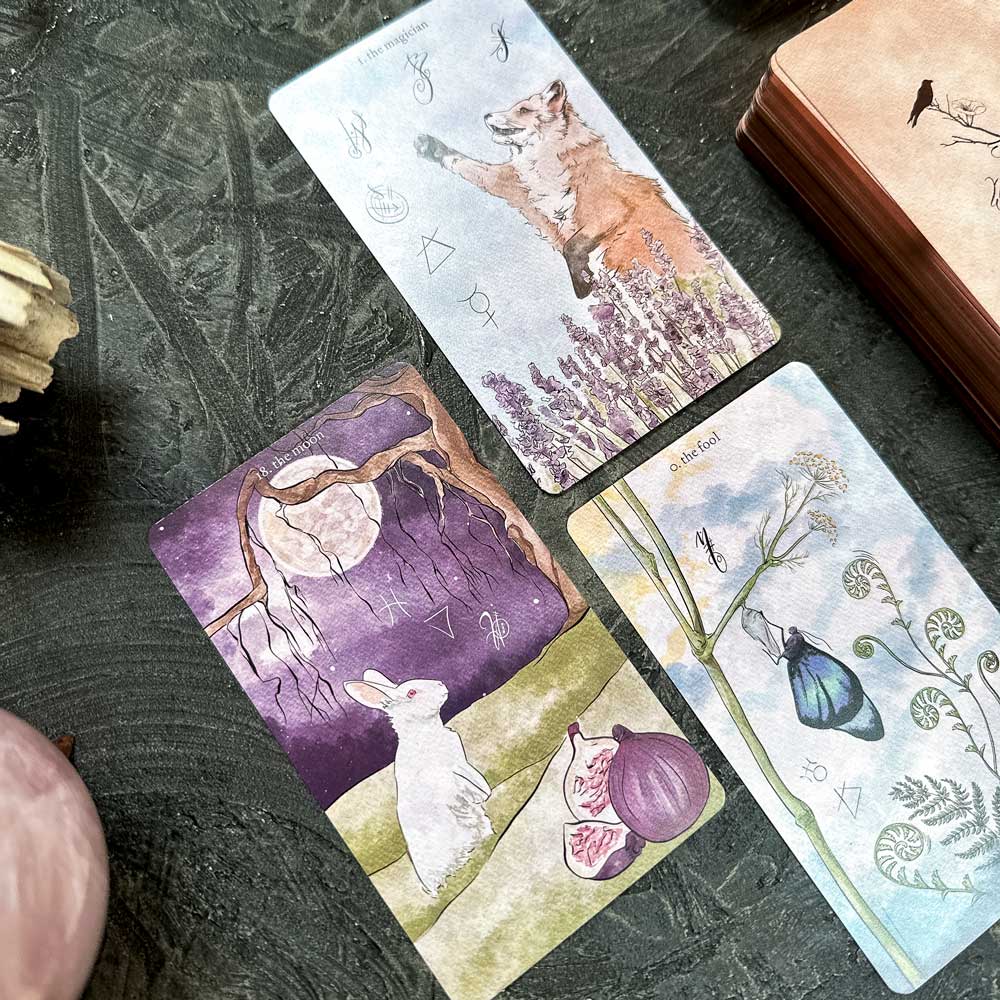 A mystical scene showcases three illustrated tarot cards from The Likely Tarot on a dark surface: one card depicts a fox with lavender, another features a rabbit with figs under the moon, and the third displays various plants and symbols. A stack of other cards peeks from behind.