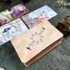 A stack of The Likely Tarot cards sits on the table, with the top card featuring botanical and bird illustrations. Other nature-themed cards are scattered around, accompanied by crystals and dried foliage nearby.