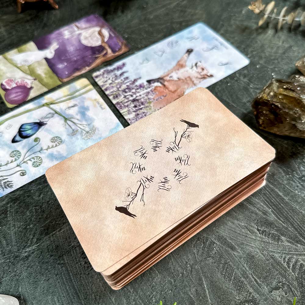 A stack of The Likely Tarot cards sits on the table, with the top card featuring botanical and bird illustrations. Other nature-themed cards are scattered around, accompanied by crystals and dried foliage nearby.