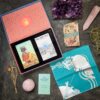 The "This Might Hurt Tarot - LE Boxed Set" includes tarot cards, an illustrated cloth, a booklet, a rose quartz sphere, a pink crystal obelisk, an amethyst cluster, and small air plants on a dark surface.
