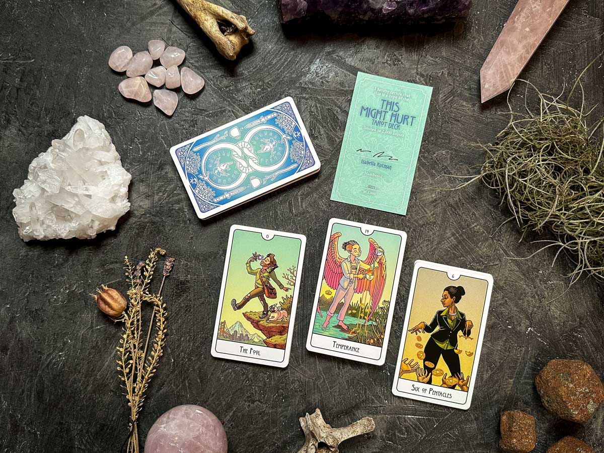 A tarot spread on a textured surface features The Fool, Temperance, and Six of Pentacles, accompanied by crystals, a bone, dried herbs, and the sleek "This Might Hurt Tarot - LE Boxed Set" box to complete the mystical setup.