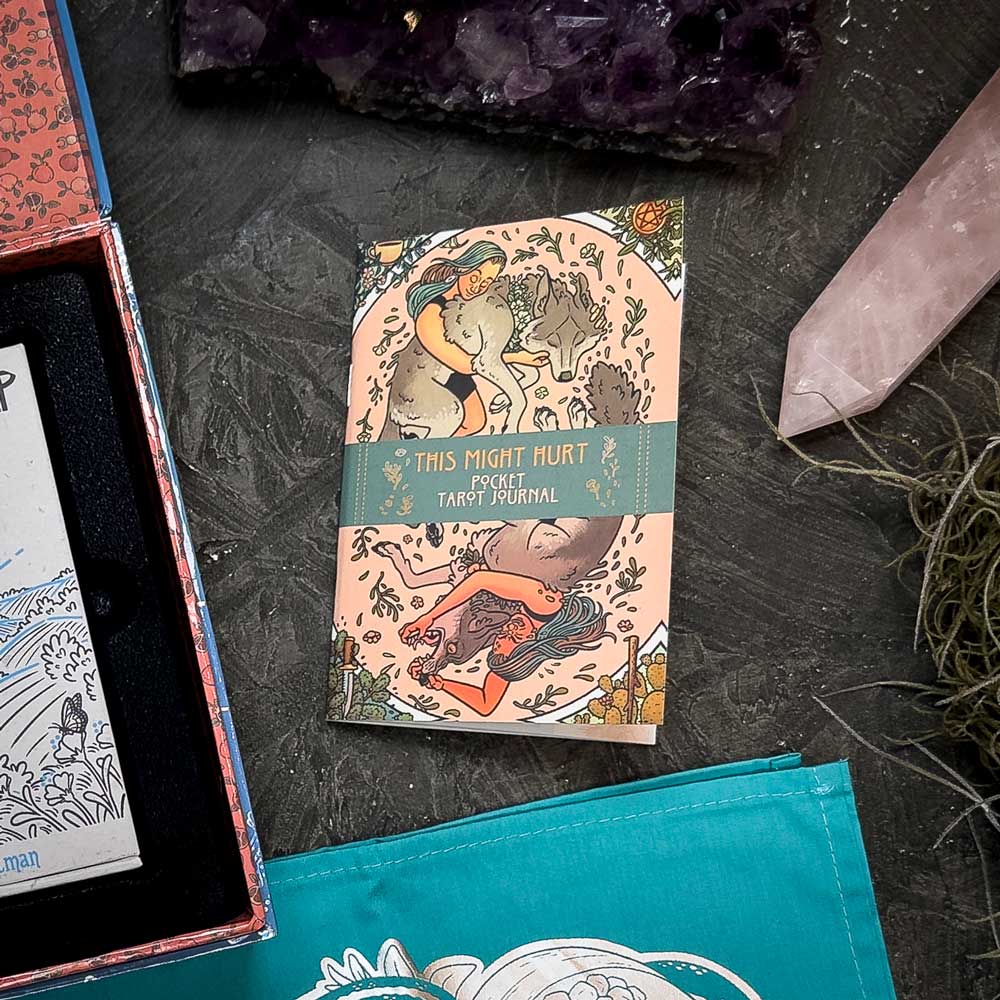 The "This Might Hurt Pocket Tarot Journal" from the "This Might Hurt Tarot - LE Boxed Set" lies on a dark surface, adorned with vibrant illustrations of a woman and animals, encircled by crystals and a blue cloth, creating an enchanting scene.
