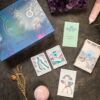 The "This Might Hurt Tarot - LE Boxed Set" contains a themed box, booklets, three illustrated cards, a dried flower, pink crystal, round stone, and purple crystal cluster on a dark textured background.