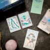 A Tarot set from This Might Hurt Tarot - LE Boxed Set lies on a textured surface with three cards, including "The Magician's Wand" and "The Middle Path." It comes with a guidebook, pink stone, packaging, and leaflets.