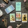 A tarot card spread features "The Fool," "Temperance," and "Six of Pentacles" on a dark surface. Nearby, the closed This Might Hurt Tarot - LE Boxed Set rests among dried plants, rose quartz, and a shimmering crystal.