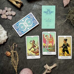 A tarot card spread features "The Fool," "Temperance," and "Six of Pentacles" on a dark surface. Nearby, the closed This Might Hurt Tarot - LE Boxed Set rests among dried plants, rose quartz, and a shimmering crystal.